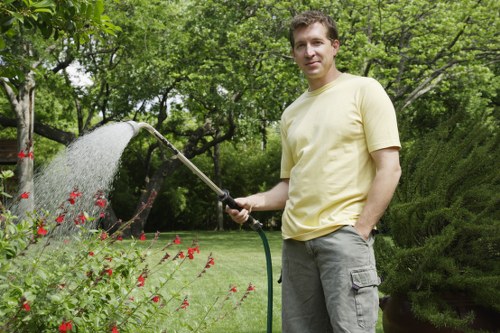 A man stands in a well-maintained garden, holding a garden hose and watering a flower bed with red blooms. The garden features a lush, green lawn with dense grass, surrounded by various shrubs and trees that create a natural privacy screen. In the background, there are taller trees with leafy canopies providing shade and an inviting outdoor environment. The scene is set on a bright day with natural sunlight illuminating the space, emphasizing the vibrant greenery and colorful flowers. The garden appears to be part of a residential outdoor space in Swiss Cottage, reflecting careful landscaping and regular garden maintenance typical of professional gardening services. The man’s casual attire, including a pale yellow t-shirt and grey cargo shorts, suggests he is engaged in routine outdoor garden tasks like watering, with the setting offering a tranquil and cultivated atmosphere ideal for gardening and lawn care support from companies like Hedge Trimming Swiss Cottage. The overall composition highlights a tidy outdoor area with an emphasis on plant health and garden aesthetics, suitable for garden service websites emphasizing outdoor maintenance in London postcodes close to Swiss Cottage.