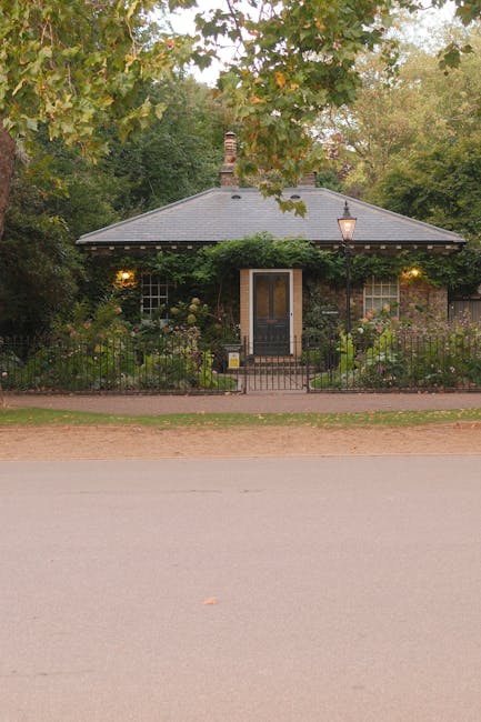 A cozy, single-story brick cottage with a dark gray, hipped roof and a brick chimney, surrounded by a neatly maintained garden. The garden features lush green shrubs, flowering plants, and a variety o